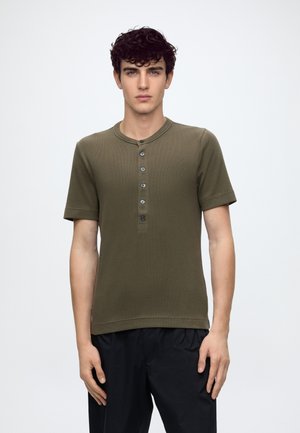Jeune homme aux cheveux bouclés foncés portant un t-shirt henley à manches courtes vert olive avec cinq boutons et un pantalon noir, debout devant un fond uni.