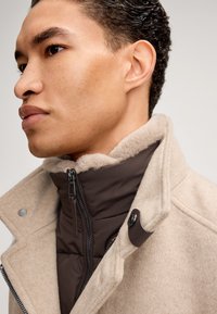 Beige wollen blend jas met een bruine gewatteerde binnenlaag. Kenmerkt zich door een hoge kraag met een zachte fleece voering en een drukknoopdetail.