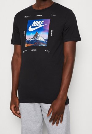Schwarzes Nike T-Shirt mit einem Bergmotiv und weißem Schriftzug "NIKE AIR" und "Just Do It", getragen von einer Person in hellgrauer Hose.