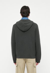 Felpa grigia con texture a coste e cappuccio regolabile con lacci. Presenta maniche lunghe e orlo dritto, abbinata a pantaloni cargo beige.