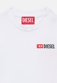 Άσπρο βαμβακερό μπλουζάκι με στρογγυλή λαιμόκοψη. Διαθέτει κόκκινο λογότυπο με τη λέξη "DIESEL" σε έντονο μαύρο κείμενο στο αριστερό στήθος. Η ετικέτα είναι έντονα κόκκινη.