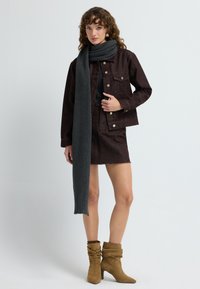 Giacca di jeans marrone con bottoni dorati, abbinata a una gonna coordinata. Una sciarpa grigia scuro a coste e stivali con tacco morbidi di colore beige completano l'outfit.