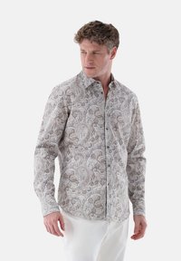 Long-sleeve button-up shirt in a beige and brown paisley pattern, featuring a spread collar and dark buttons, worn with white pants.