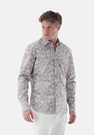 Langärmliges Hemd mit Knopfleiste in einem beigen und braunen Paisleymuster, mit einem klassischen Kragen und dunklen Knöpfen, getragen mit weißen Hosen.