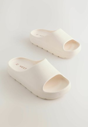 Valkoiset slip-on-sandaalit, joissa on avoin varvas ja jotka on valmistettu teksturoidusta materiaalista. Niissä on paksu pohja ja sileä, minimalistinen muotoilu.