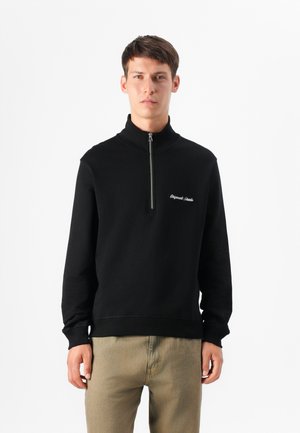 Jeune homme portant un sweat à capuche zippé noir avec un logo, se tenant contre un fond blanc, regardant directement l'objectif.