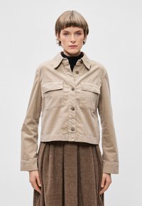 Veste cropped beige en coton, avec un col, deux poches poitrine et une fermeture à bouton à l'avant. Texture lisse et légère.
