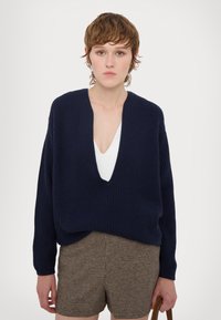 Laivansinisessä, ribbipintaiseen neulepuserossa on syvä V-kaula-aukko ja rento istuvuus. Se on pukeutunut valkoisen paidan päälle, yhdistettynä ruskeisiin houndstooth-shorteihin ja ruskeaan laukkuun.