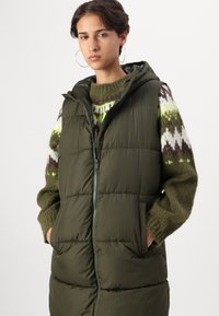 Oliven grønn puffer vest, med glidelås foran, hette og to sidelommer. Brukt over en strikket genser med et grønt og brunt geometrisk mønster.