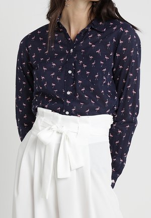 Granatowa koszula z różowym motywem flamingów wpuszczona w białe, wysokie spodnie z paskiem zawiązanym na kokardę w talii.