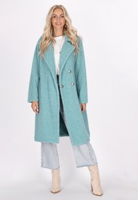 Cappotto texture verde acqua con design doppiopetto, bottoni argentati e ampi rever, indossato sopra una maglietta bianca e jeans in denim chiaro.