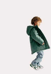Zelená péřová bunda s kapucí, přes kterou je oblečený světle modrý svetr, doplněná volnými světle modrými džíny a teniskami s zelenými akcenty.