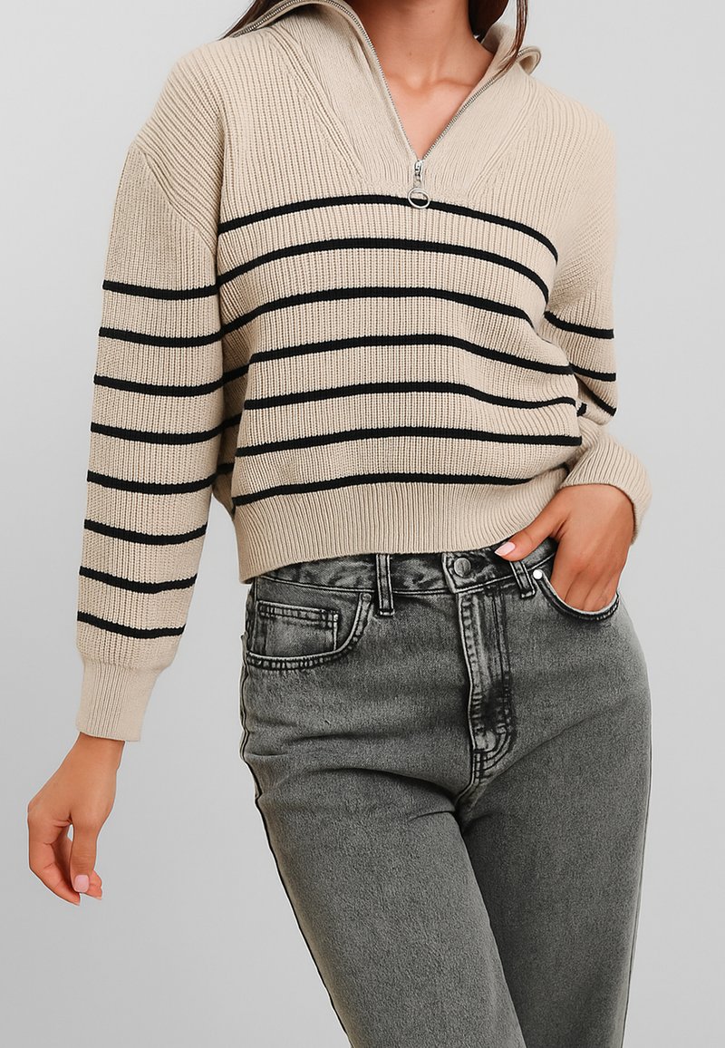 Pullover beige in maglia con zip a un quarto, caratterizzato da strisce orizzontali nere. Abbinato a jeans grigi scuri a vita alta e con texture.