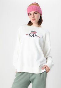 Biely sweatshirt s logom GAP a americkou vlajkou, doplnený mätovozelenými teplákmi. Modelka nosí ružový pruhovaný pletený čiapku.