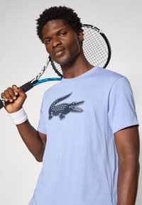 T-shirt de manga curta lilás claro com um logótipo de crocodilo preto. O modelo segura uma raquete de ténis, exibindo um visual desportivo e casual.