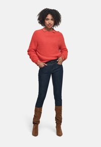 Maglione rosso peloso con colletto decorativo, jeans in denim scuro e stivali al ginocchio in suede marrone. L'outfit ha una vestibilità rilassata e dettagli testurizzati.