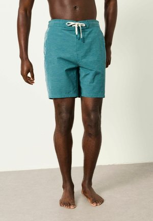 Male model wearing teal swim shorts with white drawstring and side stripes, standing barefoot on a light floor against a plain background.