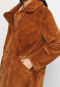 Femme portant un manteau en fausse fourrure marron pelucheux avec col et poches latérales, une main placée dans une poche.