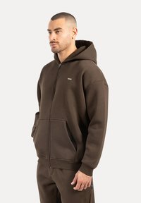 Muž s krátkými vlasy, oblečený v hnědé mikině na zip a odpovídajících kalhotách, stojící s rukama v kapsách na jednobarevném pozadí.