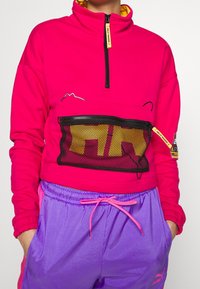 Sudadera fucsia con cremallera y bolsillo de malla, detalles en amarillo y diseño de montaña. Combinada con pantalones cortos ligeros morados con cintura elástica.