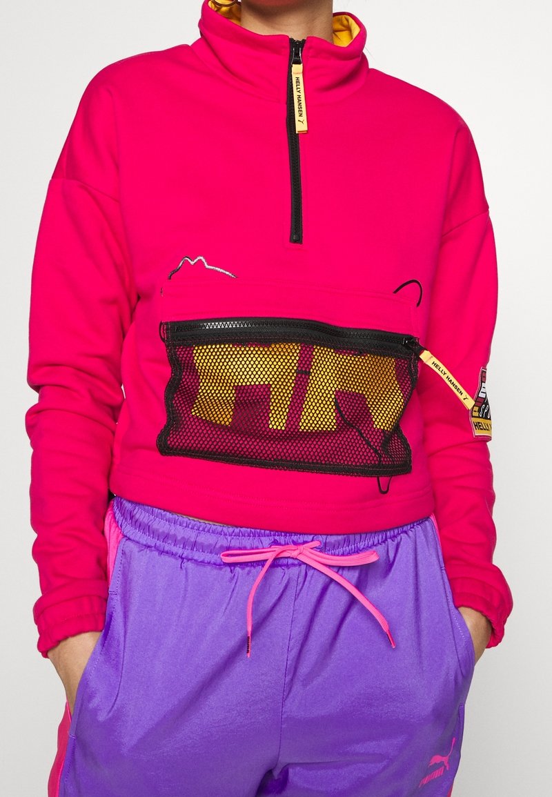 Sudadera fucsia con cremallera y bolsillo de malla, detalles en amarillo y diseño de montaña. Combinada con pantalones cortos ligeros morados con cintura elástica.
