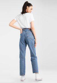 Jeans de ganga azul de cintura alta com um corte descontraído, cinco bolsos e um retângulo de couro castanho nas costas. Combinados com uma t-shirt branca simples.