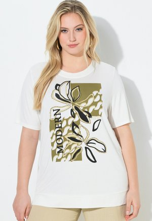 Lyshåret kvinde iført en hvid skjorte med korte ærmer med et grønt og sort abstrakt bladmotiv og teksten "MODERN", beige bukser og gyldne øreringe i hoop-design.