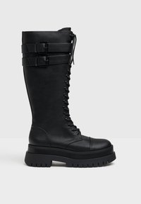 Bottes de combat en cuir noir, montantes jusqu'au genou, avec semelle épaisse, présentant un laçage à l'avant et deux sangles ajustables avec boucles.