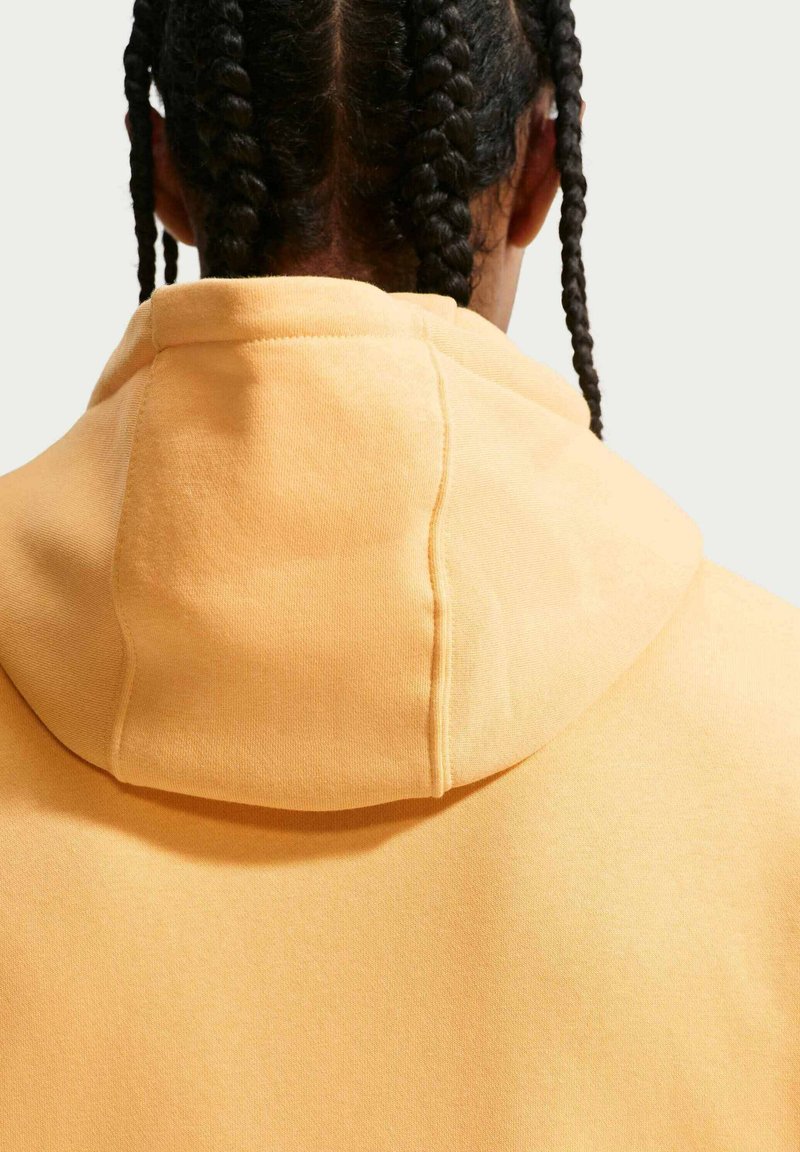 Person mit schwarzen geflochtenen Haaren, von hinten gesehen, trägt einen hellorangen Kapuzenpullover vor einfarbigem Hintergrund.