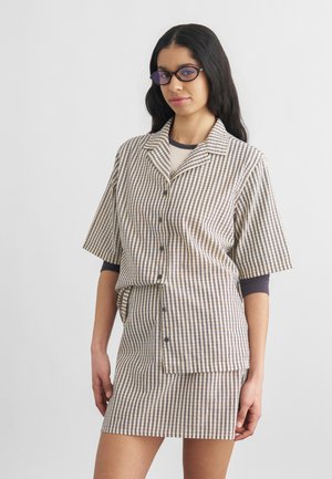 Νεαρή γυναίκα με μακριά μαύρα μαλλιά φοράει σκουρόχρωμα γυαλιά, ένα εφαρμοστό t-shirt με στρώσεις, πουκάμισο με κοντά μανίκια σε μπεζ και καφέ καρό σχέδιο με κουμπιά και φούστα που ταιριάζει.