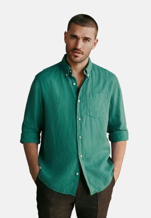 Man with short hair and beard wearing a green button-up shirt with sleeves rolled up and dark pants, hands in pockets, against white background.