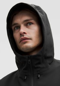 Svart vattentät jacka med hög krage och justerbar huva. Har en frontdragkedja, elastiska dragsnören och en smidig textur.