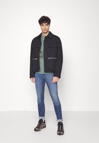 Chaqueta exterior negra con dos bolsillos frontales con cremallera, llevada sobre una camiseta verde, combinada con jeans azules y zapatillas negras con acentos naranjas.