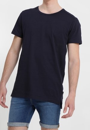 Mężczyzna w prostym czarnym t-shircie z krótkim rękawem i podwiniętych niebieskich dżinsowych szortach na białym tle.