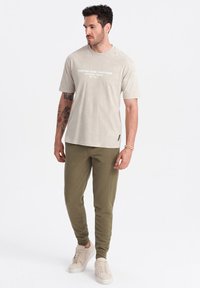 Beige t-shirt met korte mouwen en witte tekst, gecombineerd met olijfgroene tapered joggers en lichte sneakers, staande tegen een witte achtergrond.