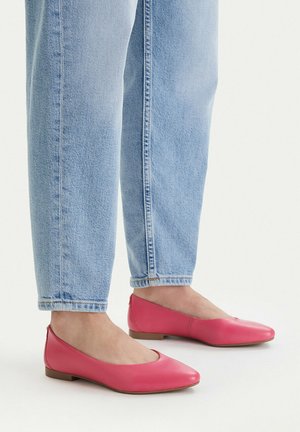 Voeten met felroze ballerina's gecombineerd met lichtblauwe spijkerbroek tegen een effen witte achtergrond.
