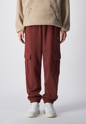 Beiger Fleece-Pullover mit Reißverschluss-Taschen und bordeauxroten Cargo-Hosen mit seitlichen Taschen. Die Jogginghosen haben elastische Bündchen. Weiße Sneaker mit Akzenten.