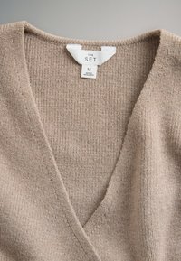 Beige geribd gebreid V-hals kledingstuk met een wit label met de tekst "THE SET", maat M, gemaakt in Bangladesh.