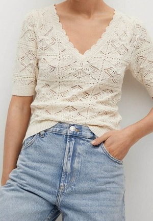 Femme portant un haut en maille crème à manches courtes avec des motifs en losange et un jean taille haute bleu clair, main dans la poche.