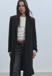 Musta villakangastakki, joka on muotoutunut huolellisesti, varustettuna lovetetulla kauluksella, yksittäisellä nappisuljenta ja sivutaskuilla, käytetty raidallisen paidan päällä.