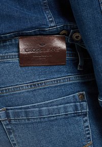 Niebieskie dżinsy z denimu z brązową skórzaną naszywką z napisem „CROSS JEANS”. Posiada widoczne szwy, detale kieszeni oraz klasyczny krój.