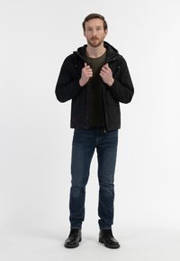 Sort zip-jakke med hætte, to forlommer, båret over en mørkegrøn skjorte og parret med blå jeans og sorte støvler.