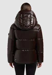 Bruine pufferjas met een glanzende afwerking, voorzien van een grote capuchon en een reliëflogo op de achterkant. Het materiaal lijkt geïsoleerd en soepel.