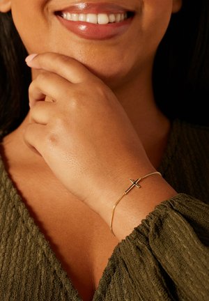 Glimlachende vrouw draagt een olijfgroen gestructureerd topje, met haar hand rustend op haar kin, met een slanke gouden armband met horizontaal kruisbedeltje om haar pols.