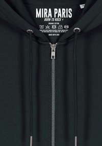 Felpa con zip di colore verde scuro realizzata in cotone biologico. Presenta una cerniera YKK, lacci e un'etichetta stampata con le istruzioni per la manutenzione.
