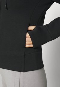 Sweat à capuche noir à texture lisse, avec une fermeture éclair à l'avant, des poches latérales et des manches ajustées. Associé à un pantalon gris clair.