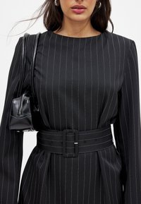 Robe noire à rayures avec des manches longues, un col rond et une ceinture avec une boucle carrée. Associée à un petit sac à main noir.