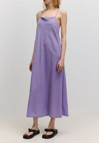 Langes lila Kleid mit dünnen Trägern, drapiertem Ausschnitt und lockerer Passform. Getragen mit schwarzen flachen Sandalen mit dicker Sohle und breiten Riemen.
