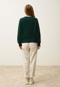 Zielony sweter dzianinowy o luźnym kroju, zestawiony z jasnobeżowymi, teksturowanymi spodniami oraz neutralnymi, masywnymi sneakersami. Minimalistyczne tło.