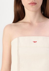 Beež rihmata top, valmistatud tekstuuriga kangast, millel on punased õmblused ja väike logo külgnes. 